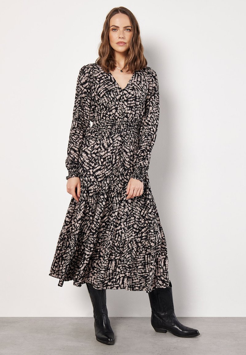 Langes, schwarz-creamfarbenes, gemustertes Kleid mit V-Ausschnitt, langen Ärmeln und gesmokter Taille. Getragen mit schwarzen Cowboys Stiefeln.