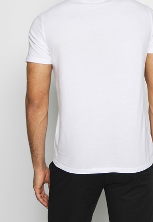 Mand iført en simpel hvid kortærmet T-shirt og sorte bukser, set bagfra med højre hånd afslappet ved siden.