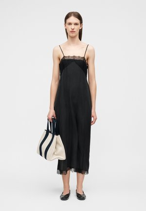 Γυναίκα με μαύρο φόρεμα με λεπτομέρειες από δαντέλα, κρατώντας μια μπεζ τσάντα tote με μαύρα λουριά, φορώντας μαύρες μπαλαρίνες, όρθια σε απλό φόντο.