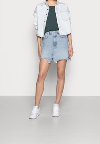 Ljusblå denimkjol med fransad nederkant, tillsammans med en grön topp och en ljus denimjacka. Vita sneakers med rosa detaljer fulländar outfiten.