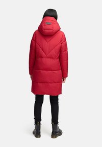 Person wearing a long red padded coat with a hood, black pants, and black lace-up boots, standing facing away on a plain background.
