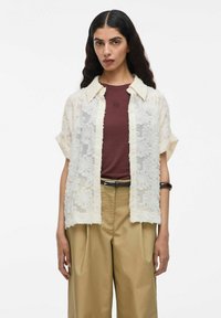 Camisa de manga corta con superposición de encaje en color crema, con cuello extendido y cierre de botones. Debajo, una camisa ajustada en color burdeos y pantalones beiges.