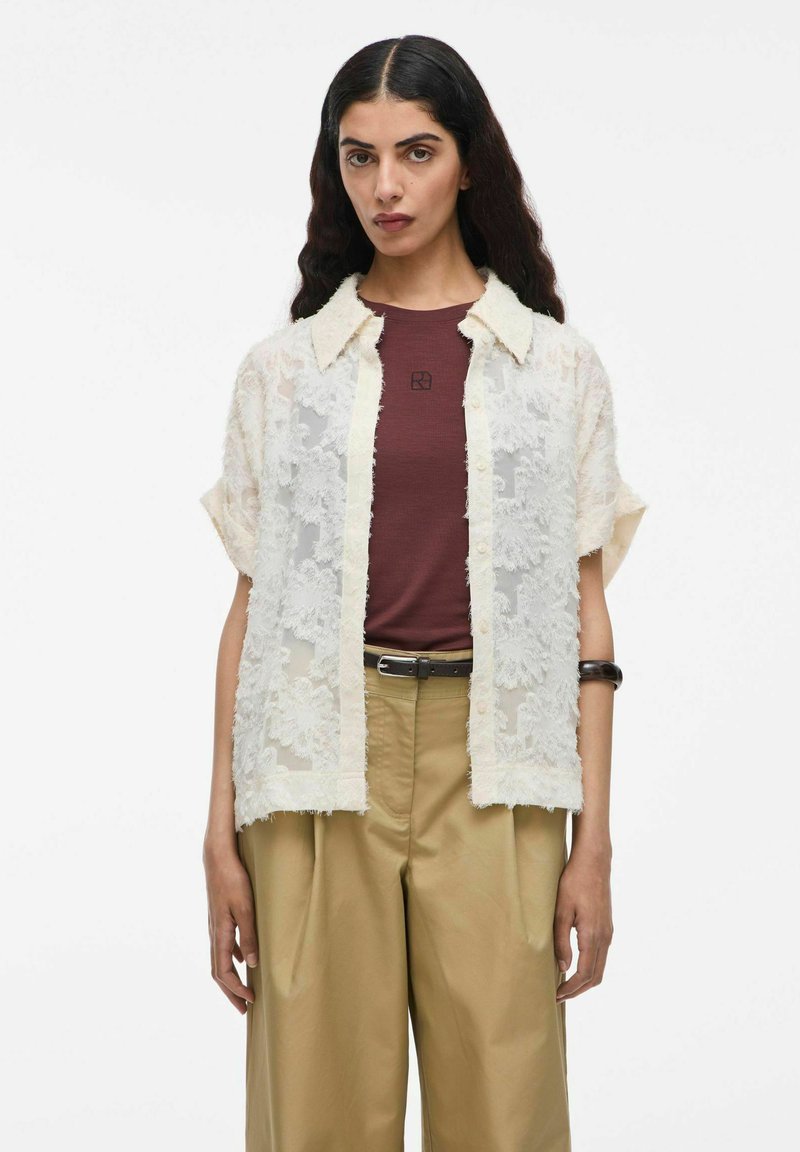 Chemise à manches courtes avec empiècements en dentelle en crème, avec un col évasé et une fermeture à boutons. En dessous, une chemise ajustée bordeaux et un pantalon beige.