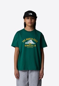 Green cotton t-shirt with yellow graphics reading "THE NORTH FACE," a mountain, and "BERKELEY, CA." Paired with gray sweatpants and a black cap.