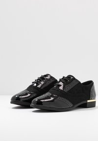 Chaussures oxford noires en cuir verni et en daim, dotées de détails perforés, d'un laçage et d'un petit accent doré sur le talon.