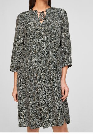 Kvinde iført en knælængde kjole med lange ærmer i sort-hvidt abstrakt prikmønster og bindebånd ved halsen.