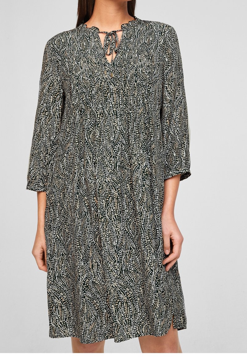Femme portant une robe mi-longue à manches longues avec un motif abstrait en points noirs et blancs et un col à nouer.