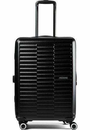 Schwarzer Hartschalenkoffer mit horizontalen Rillen, ausgezogenem Teleskopgriff, vier drehbaren Rollen und American Tourister-Logo vorne.