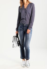 Blouse à boutons grise à manches longues, jeans bleus avec des bords effilochés, et baskets argent métallisé. Tenant un sac en argent brillant.