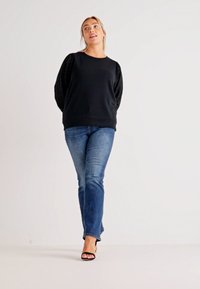 Black sweatshirt with textured sleeves, paired with blue flared jeans. The model is wearing black high-heeled sandals. Smooth fabric, relaxed fit.