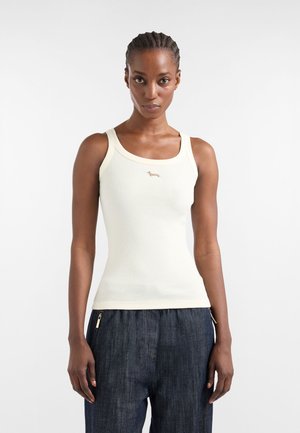 Donna con capelli intrecciati che indossa un top smanicato aderente color avorio chiaro a coste e pantaloni larghi scuri, su uno sfondo chiaro e uniforme.