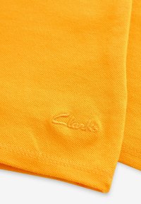 Oranžová bavlněná látka s texturovaným povrchem, která má na jednom okraji vyšité logo "Clark" v světlejší oranžové barvě.