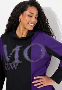 Svart och lila sweatshirt med kapuschong, med stort "MO"-design prytt med rhinestones och en mjuk textur. Långa ärmar, avslappnad passform.