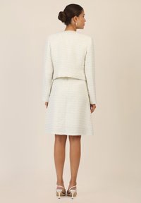 White textured two-piece suit with a cropped jacket and knee-length skirt, featuring a subtle sheen and embellishments at the collar.