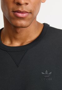 Musthave musta tekstureeritud dressipluus ümmarguse kaelusega ja keskosas diskreetne tikitud Adidas'i logo. Materjal näib olevat pehme ja hingav.