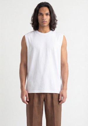 Giovane uomo con capelli scuri di media lunghezza, che indossa una camicia bianca senza maniche e pantaloni marroni, di fronte a uno sfondo semplice.