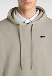 Beige huvtröja med dragsnören, prydd med en liten svart skateboard-logotyp på framsidan och lagd över en vit skjorta.