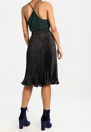 Jupe plissée noire tombant au mi-mollet, associée à un haut vert avec un dos drapé. Le mannequin porte des bottes à talons en velours bleu.