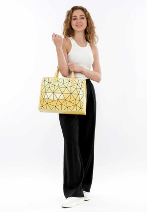 Geel geometrische tote bag met een driehoekig patroon in zwart op een glanzend oppervlak, met stevige handvatten. Draagt bij zwarte broeken en sneakers.