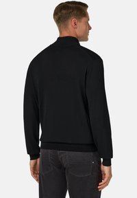 Melnā adītā džemperis ar augstu apkakli un ribotām piedurknēm. Gluda tekstūra, pieguļošs siluets un minimālisma dizains. Parādīta aizmugures skata.