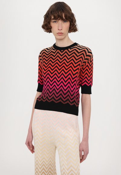 Pull en tricot avec un motif en chevrons dans des dégradés de rouge, rose et noir ; col et manches côtelés ; associé à un pantalon clair à motifs.