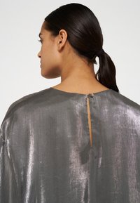 Grau metallic Bluse mit einem Schlitz hinten und einem Knopfverschluss. Der Stoff hat eine glatte, glänzende Textur und ein locker sitzendes Design.
