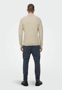 Hellbeige gerippter Pullover mit hohem Kragen, kombiniert mit dunkel karierten Hosen mit blauen und roten Akzenten. Schwarze Schuhe vervollständigen den Look.