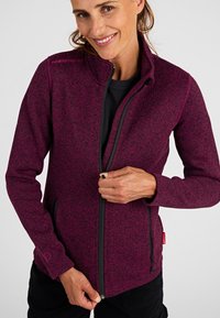 Una mujer sonriente cerrándose una chaqueta de cremallera color marrón sobre una camiseta negra frente a un fondo liso.