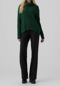 Mujer vistiendo un suéter de cuello alto de canalé verde oscuro, pantalones de pierna ancha negros y zapatos de plataforma negros, de pie sobre un fondo gris.