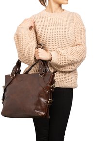 Borsa tote in pelle marrone con grande corpo, doppie maniglie e dettagli in metallo dorato. Indossata con un maglione beige testurizzato e pantaloni neri.