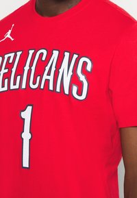 Red cotton t-shirt featuring "PELICANS" in white and gray text and the number "1" in white. Includes a small logo on the left shoulder.
