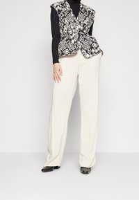 Svart och vitmönstrad väst med draperad front, bärs över en svart polotröja och krämfärgade vida byxor. Slät textur och skräddarsydd passform.