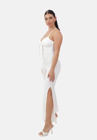 Vestido blanco ajustado con tirantes finos, un lazo atado en el frente, aberturas en los costados y tejido texturizado. Llevado con sandalias blancas de tacón.