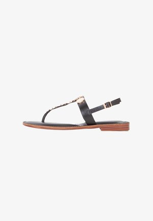 Sandal de cuero negro plano con diseño de tira en T, herrajes de acento dorado, suela de goma marrón y correa ajustable en el tobillo. Textura suave.