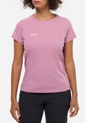 Femme portant un t-shirt de sport rose à manches courtes avec le logo "MILLET" et un pantalon noir, se tenant devant un arrière-plan uni.