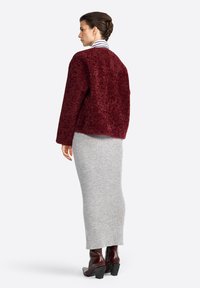 Femme de dos, portant une veste en fausse fourrure bordeaux, une jupe midi gris côtelé et des bottines à talons épais noires, sur fond uni.