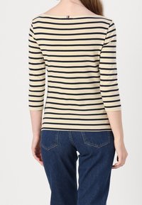Pruhované tričko v krémové a námořnické barvě, s rukávy po tři čtvrtiny, kulatým výstřihem a malým detailem smyčky na zadní straně. V kombinaci s modrými džínami.