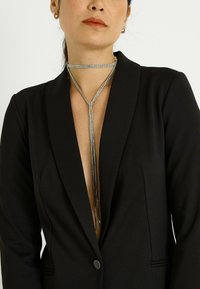 Collier ras-de-cou en strass argentés avec un design lariat, comprenant une pendaison centrale qui s’étend vers le bas, porté sur une veste blazer noire.