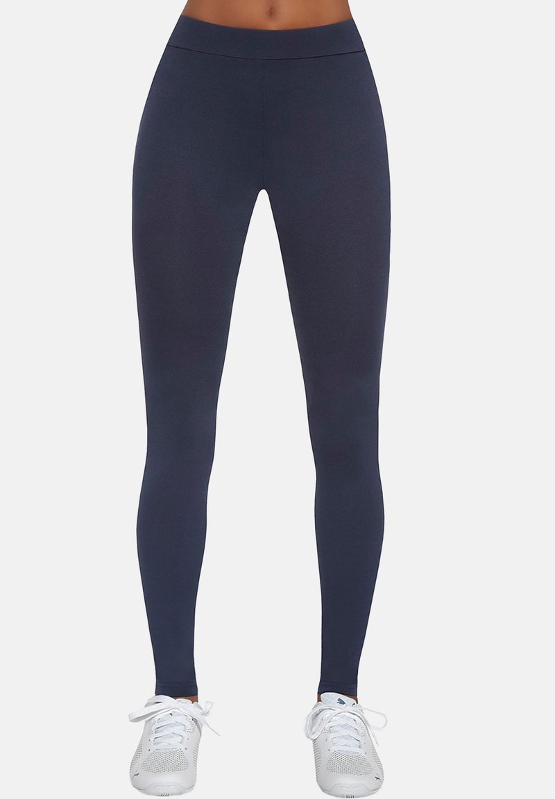 Granatowe legginsy sportowe z gładkiego, elastycznego materiału. Wysoki pas i dopasowany krój. Połączone z białymi trampkami na neutralnym tle.
