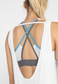 Top bianco senza maniche con schiena scoperta, che rivela un reggiseno sportivo a fasce grigio e blu sottostante. Tessuto leggero, vestibilità rilassata e spalline incrociate.