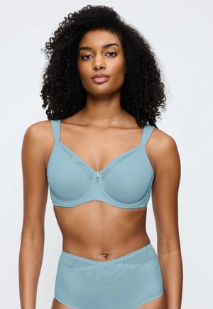 Woman with curly hair wearing a light blue supportive bra and matching high-waisted underwear, standing against a plain background.