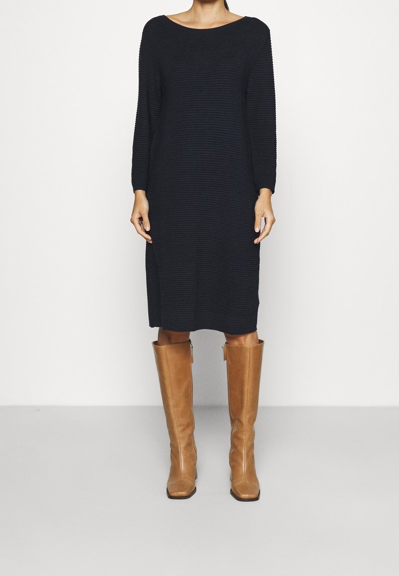Femme portant une robe midi noire côtelée à manches longues et des bottes en cuir marron montant jusqu'aux genoux, debout devant un fond uni et clair.