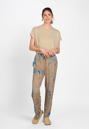 Vrouw die een beige top met korte mouwen en beige bloemenbroek draagt, staat met samengevouwen handen en glimlacht, tegen een witte achtergrond.