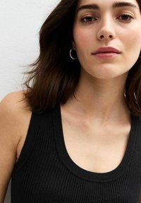 Svart ribbad tanktop med rund halsringning. Modellen har medellång brunt hår och bär silverfärgade hängehängen. Slät hudstruktur.