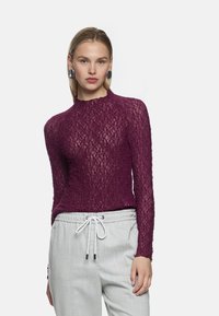 Bordeauxfarbenes Strickoberteil mit langen Ärmeln, strukturiertem Muster, hohem Kragen und figurbetontem Schnitt, kombiniert mit hellgrauen Jogginghosen mit Kordelzug.