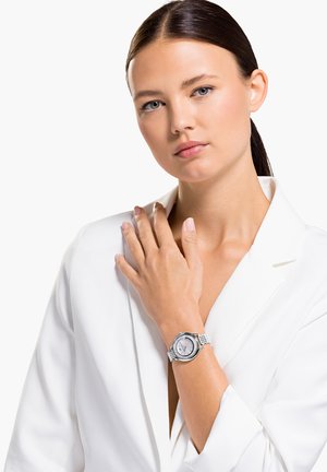 Silberne Armbanduhr mit rundem Ziffernblatt, strukturiertem Armband und weißem Ziffernblatt. Das Modell trägt ein weißes Blazer und präsentiert die Uhr an ihrem Handgelenk.