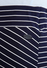 Mörkblått tyg med vita horisontella ränder, med en liten oval svart etikett som läser "Denim" fäst på sidan.