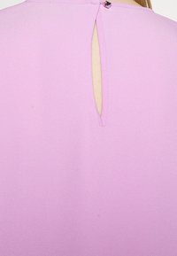 Blouse rose à la texture douce et lisse, avec une ouverture en forme de trou de serrure dans le dos, maintenue par une petite attache métallique.