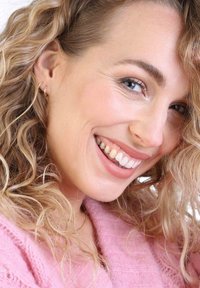 Cheveux blonds bouclés, portant des boucles d'oreilles en or, souriant avec les dents visibles. Un pull tricoté rose pâle est partiellement visible dans le cadre.