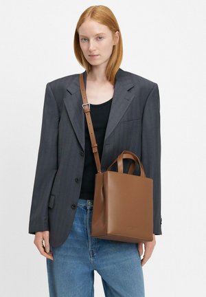 Brązowa skórzana torba tote o wyraźnym kształcie, z krótkimi i długimi uchwytami oraz minimalistycznym designem, noszona z szarym żakietem i dżinsami.